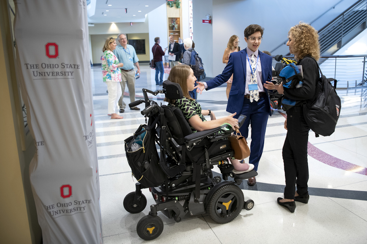 A young man speaks with two young women, one is standing and one sits in a motorized wheelchair, in an atrium in the Ohio Union.
