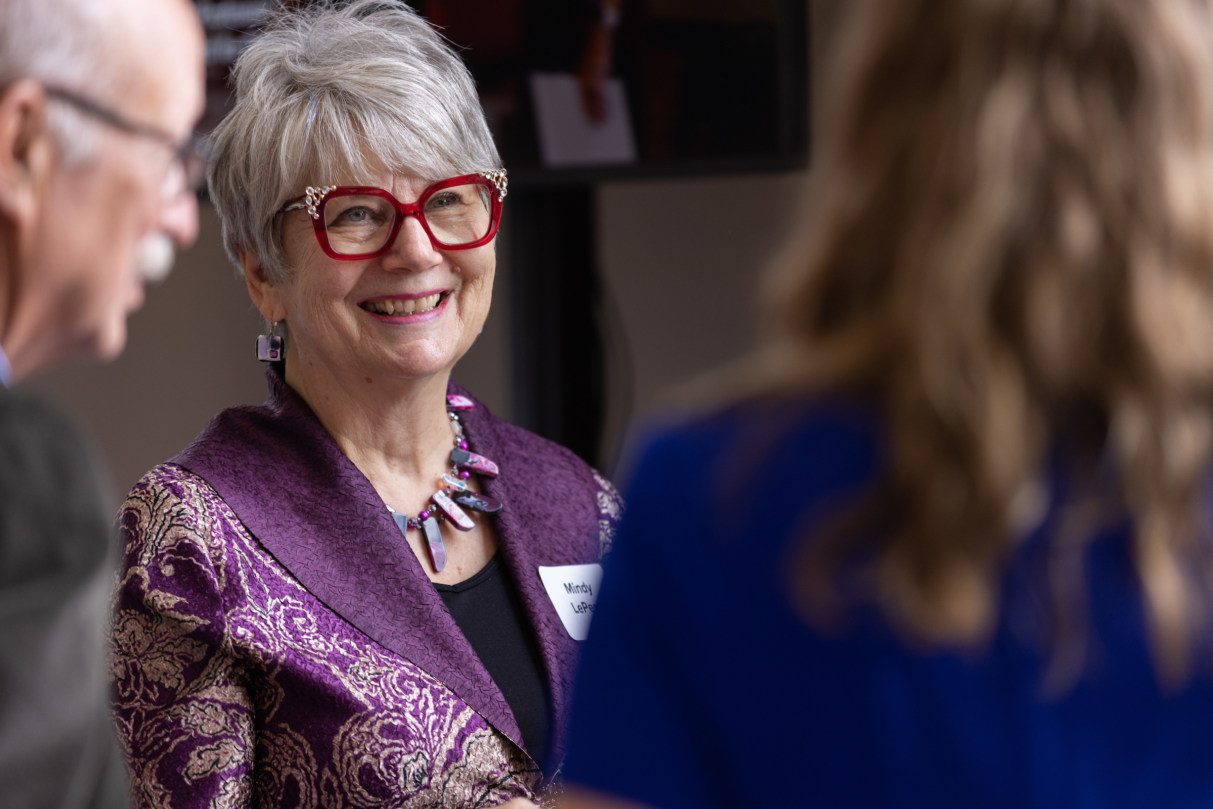 A woman faculty member with red glasses speaks to colleagues at the awards ceremony reception.