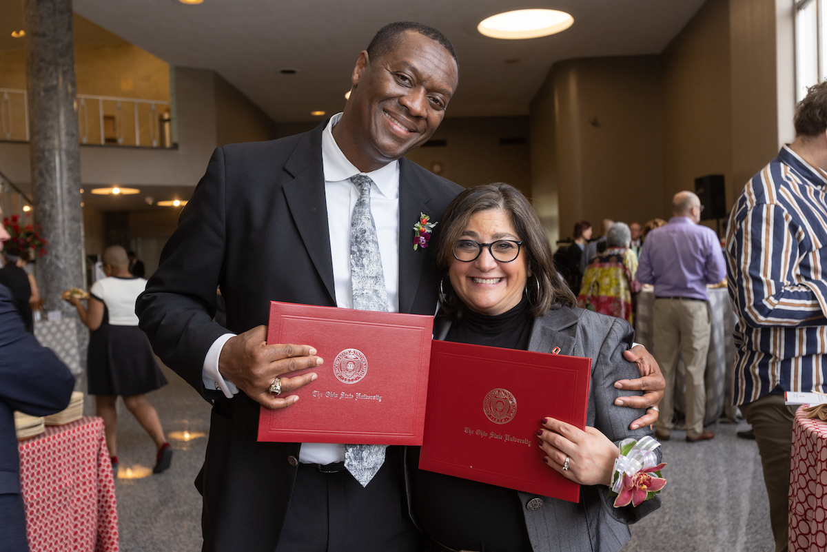 Two faculty members embrace as they show off their awards to the camera.