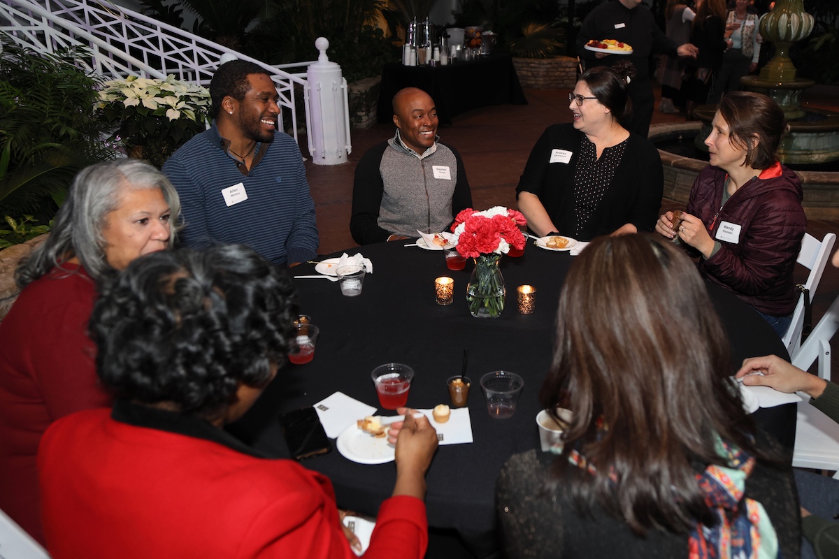 Large group of people sit at a round table sharing a conversation and smiling