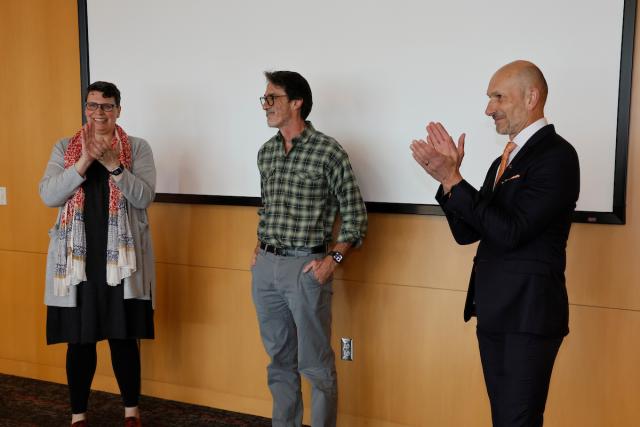 Michael LIsa stands in a classroom while two university leaders applaud.