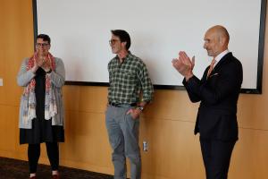 Michael LIsa stands in a classroom while two university leaders applaud.